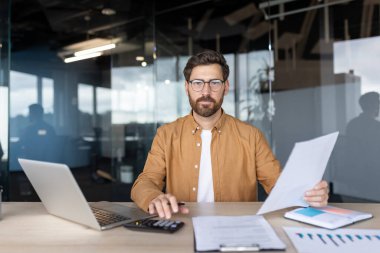 Modern ofis ortamında çalışan gözlüklü profesyonel bir birey, belgeleri analiz ediyor. Çalışma alanları, üretkenliği ve odaklanmayı yansıtan bir dizüstü bilgisayar, hesap makinesi ve organize materyalleri içerir.