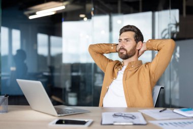Profesyonel erkek, bir iş yerinde oturmuş, arkasına yaslanıyor, üretkenlik ve düşünme ile sakin bir atmosfer sergiliyor, bir dizüstü bilgisayar, belgeler de dahil olmak üzere.