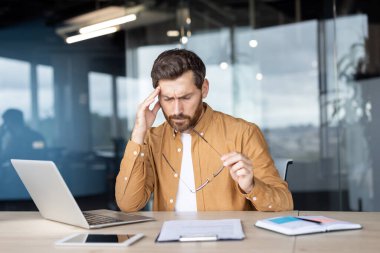 Düşünceli bir işadamı modern ofis ortamında bir masada oturuyor, belgeleri inceliyor ve bardakları tutuyor, problem çözme veya karar verme anlarında odak ve düşünce paylaşımı yapıyor..