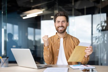 Modern ofis sahnesinde elinde bir zarf tutan gülümseyen bir adam coşkulu bir kutlama yapıyor. Profesyonel ve canlı bir çalışma ortamında başarı, başarı ve olumlu haberleri temsil eder.