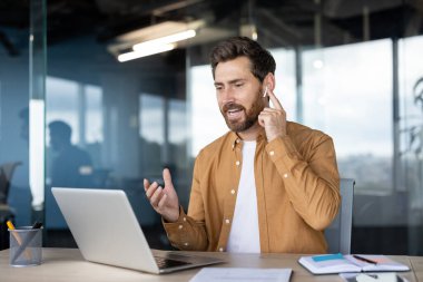 Profesyonel bir adam, ofiste kulaklık ve laptop kullanarak video konferans görüşmesi yapıyor. İşbirlikçi ve üretken bir çevreyle yaptığı konuşma sırasında meşgul ve odaklanmış görünüyor..