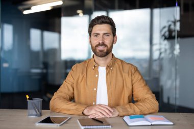 İyi ışıklandırılmış bir kurumsal ofiste masada oturan gülümseyen bir adamın portresi. Masasında defterler ve elektronik cihazlar var. Profesyonel ve ulaşılabilir bir atmosfer yaratıyor..
