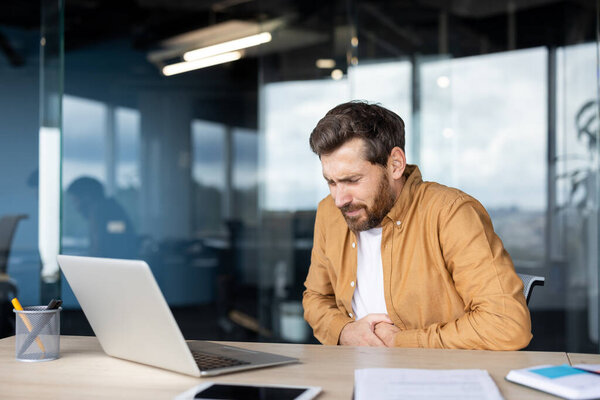 A man in casual clothing sitting at a desk holding his stomach due to discomfort while working on his laptop in a modern office setting.