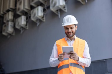 Kasketli ve yansıtıcı yelekli gülümseyen bir inşaat işçisi tabletle çalışır. Kişi dışarıda, monte edilmiş havalandırma ünitelerinin altında modern çalışma akışını gösteriyor.