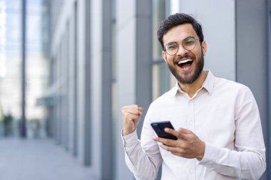 Heyecanlı iş adamı, elinde telefon tutarak başarıyı, başarıyı ve olumluluğu sembolize ediyor.