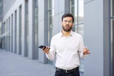 Genç bir profesyonel, elinde akıllı telefon ve kredi kartı tutarken karışık duygular sergiliyor. Modern kentsel zemin, iş ve mali karar verme dinamikleri bağlamına ekleniyor