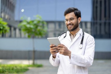 Beyaz önlük ve steteskop giyen gülümseyen bir doktor açık hava tabletinde verileri okuyarak, modern teknolojinin sağlık ortamında kendini adamış ve profesyonel olarak kullanıldığını gösteriyor..