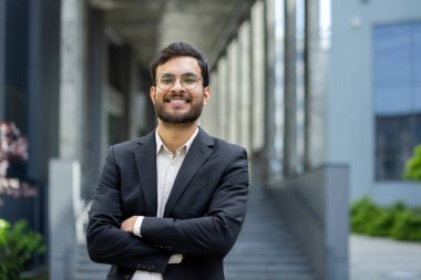 Takım elbiseli ve gözlüklü gülümseyen bir işadamı açık şehir ortamında rahatça poz verir..