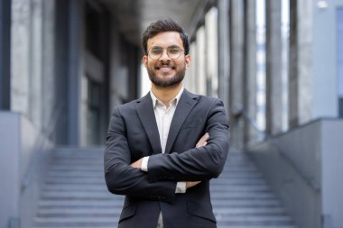 Profesyonel genç işadamı, kollarını kavuşturup, başarısını ve hırsını sergileyerek kentteki bir ortamda güvenle gülümsüyor. Şahıs resmi iş kıyafeti giyiyor.