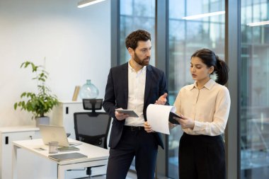 İki meslektaş, işbirliği ve takım çalışmasını vurgulayan parlak ve modern ofis ortamında iş belgelerini tartışıyor.
