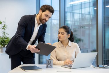Çağdaş bir ofiste, iletişim ve verimlilik üzerine yoğunlaşan, dizüstü bilgisayarlı ve belgeli bir proje üzerinde çalışan iki iş arkadaşı.