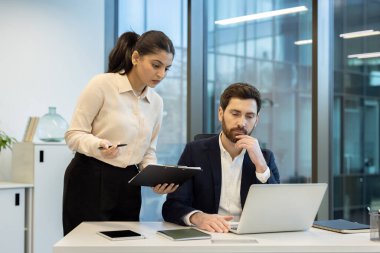 Modern bir ofiste birlikte çalışan iki meslektaşı gösteren profesyonel bir sahne. Biri bir dizüstü bilgisayar kullanarak masada otururken, diğeri bir panoya yardım ve referans notları sunar.
