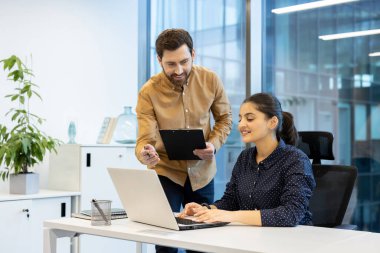 Çağdaş ofis ortamında dizüstü bilgisayar kullanan bir çalışanla konuşan bir yönetici. Takım çalışmasını ve etkili iletişimi vurgulayan rahat ve profesyonel bir atmosfer.