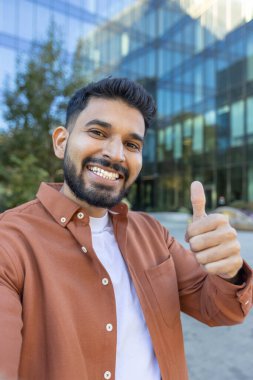 Kahverengi tişört giyen neşeli bir adam dışarıdayken baş parmak işareti yapıyor. Arka plandaki modern cam bina, pozitiflik imajını geliştiren çağdaş bir kentsel his ekliyor.