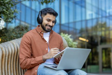 Bir adam dışarıda dizüstü bilgisayar ve kulaklık kullanarak oturuyor, gülümsüyor ve görevlere odaklanıyor..