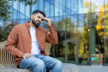 Bir adam bankta dinleniyor, başını tutuyor, yorgunluk ya da strese işaret ediyor. Konumda, kentsel yaşamı ve zorluklarını tasvir eden modern mimari ve yeşillik yer alıyor..