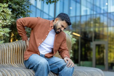 Dışarıda, tahta bir bankta oturan bir adam sırt ağrısı nedeniyle rahatsızlık gösteriyordu. Dekor, acının ve endişenin bir kombinasyonunu içeren modern bir bina ve yeşillik içeriyor.