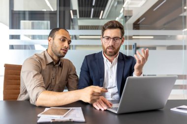 İki meslektaş bir masada oturmuş bir dizüstü bilgisayardaki bilgileri birlikte gözden geçiriyorlar. Tartışmaları, kurumsal ofis ortamında takım çalışmasının vurgulandığı odaklanmış ve profesyonelce görünüyor..
