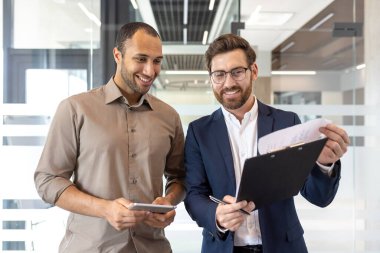 İki iş arkadaşı, modern bir çalışma alanı içinde takım çalışması, iletişim ve üretkenliği vurgulayan, bir ofis ortamında işbirliği yapan belgeler ve cihazlar kullanarak profesyonel bir tartışmaya giriştiler.