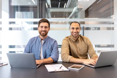 İki meslektaş, parlak ve çağdaş bir çalışma alanında dizüstü bilgisayarlarla yan yana oturuyor, gülümsüyor ve ilgileniyorlar, profesyonel bir ortamda işbirliği ve takım çalışması sergiliyorlar..