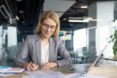 Modern bir ofisteki iş kadını elinde dizüstü bilgisayarla masa başında belgeler üzerinde çalışıyor. İş yerinde profesyonelliği, odaklanmayı ve üretkenliği temsil eden, güveni ileten