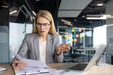 Takım elbiseli bir ofis çalışanı masasında belgeleri incelerken kafa karışıklığı gösteriyor..