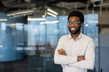 Gülümseyen profesyonel erkek, cam duvarları olan modern ofis ortamında güvenle duruyor, pozitiflik ve odak yayıyor, kariyer başarısının ve kişisel motivasyonun özünü yakalıyor..