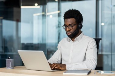 Kulaklık takan odaklanmış bir profesyonel modern ofis ortamında bilgisayarda çalışır, iletişimi, verimliliği ve teknolojiyi ön plana çıkarır..