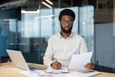 Şirket ortamında dizüstü bilgisayar üzerinde çalışırken belgelere odaklanmış bir adam.