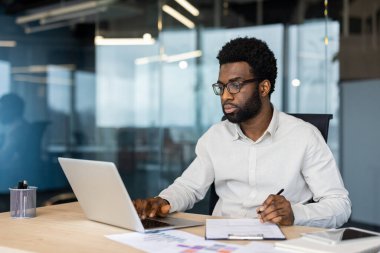 Modern ofis ortamında bir iş adamı dizüstü bilgisayar üzerinde çalışıyor ve belgeleri inceliyor, profesyonelliği, verimliliği ve kurumsal bir ortamda odaklanmayı temsil ediyor..