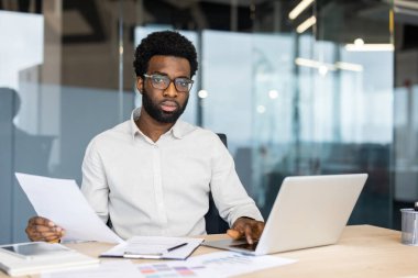 Çağdaş ofis ortamındaki bir masada dizüstü bilgisayar üzerinde çalışırken profesyonel inceleme kağıtlarına odaklanmış. Arka planda modern cam bir iç mekan var.