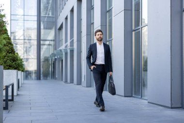 Koyu renk takım elbiseli profesyonel bir adam, modern cam cephede, güvenli bir şekilde dışarı çıkar..
