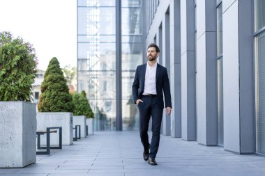 Takım elbiseli kendine güvenen bir adam yeşilliklerle çevrili bir ofis kompleksinden geçer..