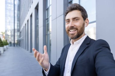 Takım elbiseli bir işadamı bir video çağrısına katılıyor, kentsel bir ortamda pozitifliğini ve bağlılığını ifade ediyor. Modern işbirliği ve profesyonel iletişimi yansıtıyor.