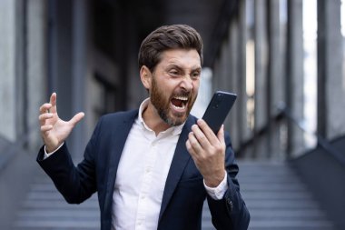 Profesyonel giyimli bir adam şehir ortamında akıllı telefonuna düş kırıklığı içinde bağırıyor. Yüzünde derin bir öfke ya da sıkıntı var.