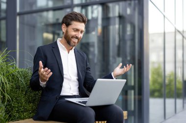 Gülümseyen bir iş adamı dizüstü bilgisayarıyla dışarıda video görüşmesi yaparken neşeli bir şekilde el kol hareketi yapıyor.