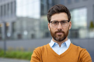 Gözlük ve süveter takan, modern ofis geçmişine karşı açık havada poz veren, profesyonellik ve duruş sergileyen iş dünyası, şirket ve liderlik teması için ideal bir adam..