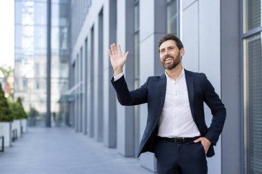 Kendine güvenen işadamı gülümseyip el sallarken modern bir şehir ortamında duruyor..