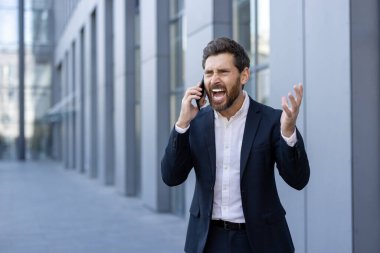 Resmi takım elbiseli bir işadamı, ofisin yakınındaki bir telefon görüşmesinde öfkeyle tepki verir. Duyguları hayal kırıklığını gösteriyor, profesyonel bir ortamda çatışmayı vurguluyor.
