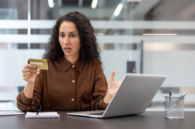 Endişeli bir kadın ofis masasında laptopuyla çalışırken kredi kartını inceliyor..