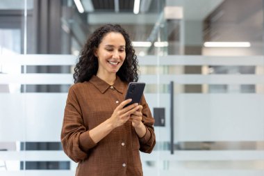 Parlak, modern ofis ortamında cam bölmeli akıllı telefon kullanan mutlu bir kadın. Sahne, iletişim, teknoloji kullanımı ve muhtemel profesyonel faaliyetler öneriyor..