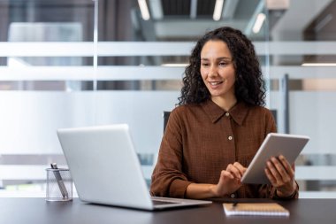 Ofis masasında oturmuş laptopla ve tabletle çalışan kendinden emin biri. Modern, organize çalışma alanı profesyonel atmosferi geliştirir, çoklu görev için teknoloji kullanımını gösterir