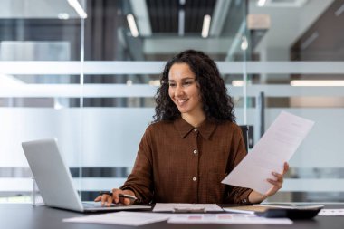 Ofiste dizüstü bilgisayarla çalışan ve belgeleri analiz eden, modern bir şirket ortamında işyeri verimliliğini ve profesyonelliğini gösteren kendine güvenen bir kadın..
