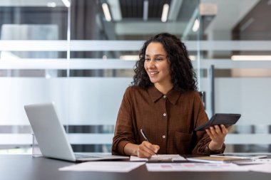 Profesyonel ofis ortamında dizüstü bilgisayar ve hesap makinesiyle çalışan gülümseyen kadın..