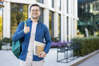 Kendine güvenen genç bir adam dışarıda duran bir defter ve sırt çantası tutuyor. Çağdaş bir çevrede başarı ve pozitifliği destekleyen neşeli bir jest..