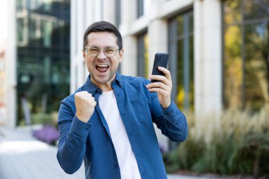 Açık havada elinde telefon tutan neşeli bir adamın görüntüsü, heyecan ve zafer ifade ediyor..