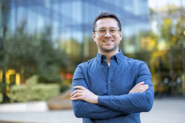 Mavi gömlekli ve gözlüklü neşeli bir adam, dışarıda güvenle dikiliyor..