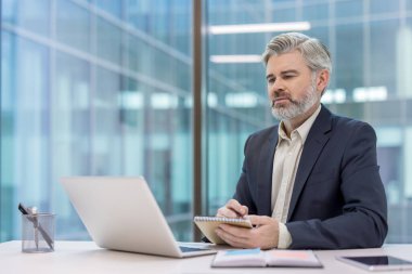 Düzgün bir şirket ortamında dizüstü bilgisayar ve defterle çalışan olgun bir iş adamı..