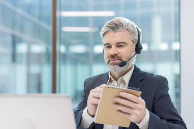 Kulaklık takan profesyonel bir adam, ofiste çalışıyor, bilgisayar başında notlar alıyor..