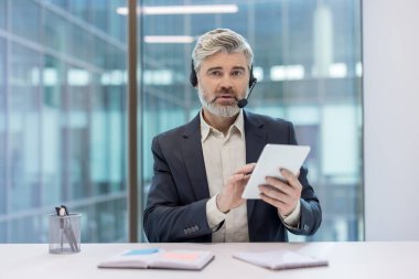 Modern ofis ortamında bir tabletle etkileşime geçen kulaklık takan bir iş adamı..